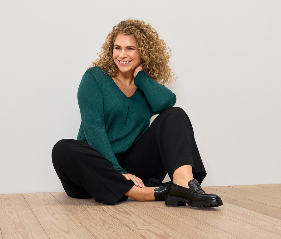 Žena sedí na podlaze v zeleném svetru a černých kalhotech.