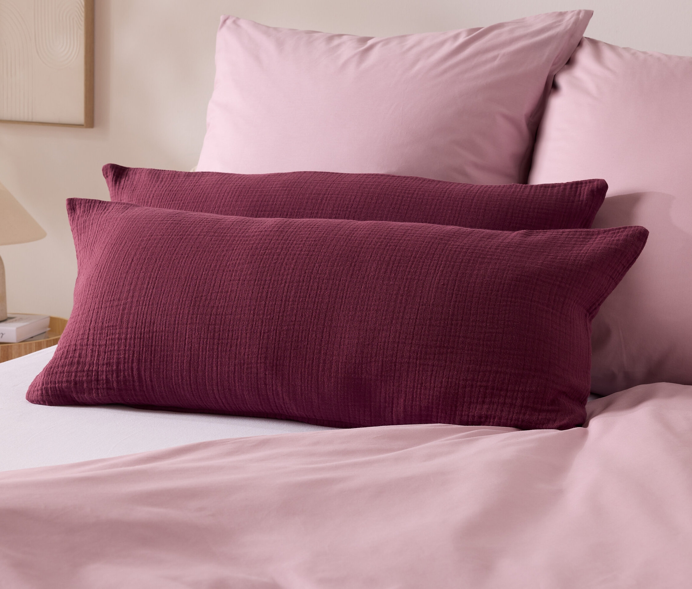 Dva bordó polštáře leží na posteli s růžovým povlečením. V pozadí jsou dva růžové polštáře a lampa.