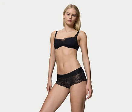 Blondýnka stojí, oblečená v černé krajkové podprsence a černých bokových kalhotkách Triumph Body Makeup Illusion Lace.