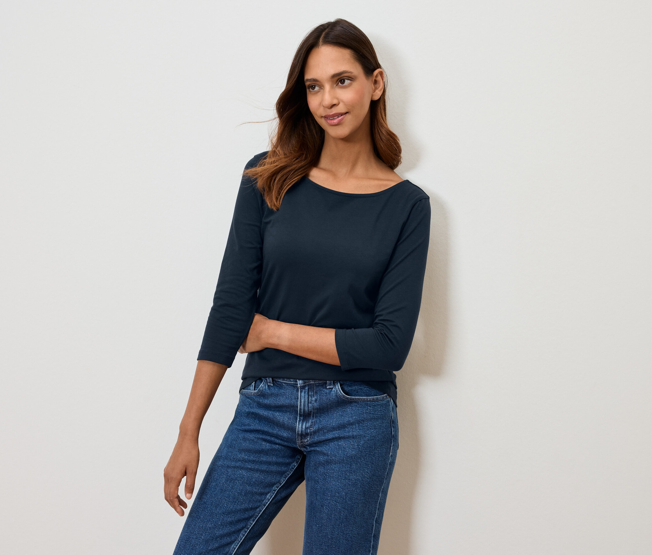 Žena pózuje v tmavě modrém tričku s tříčtvrtečním rukávem a modrých džínách.