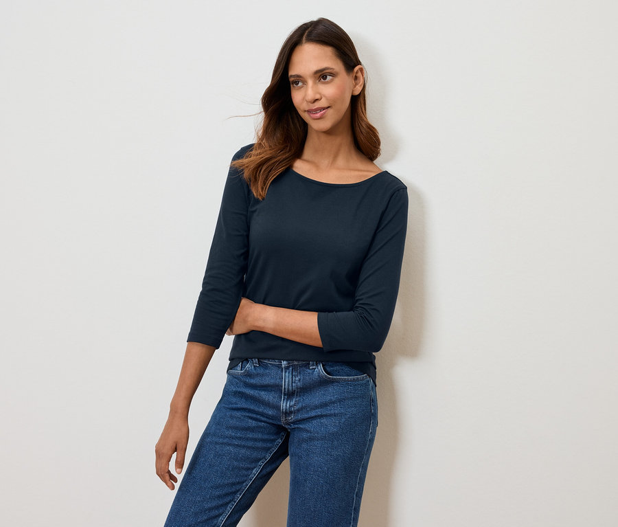 Žena pózuje v tmavě modrém tričku s tříčtvrtečním rukávem a modrých džínách.