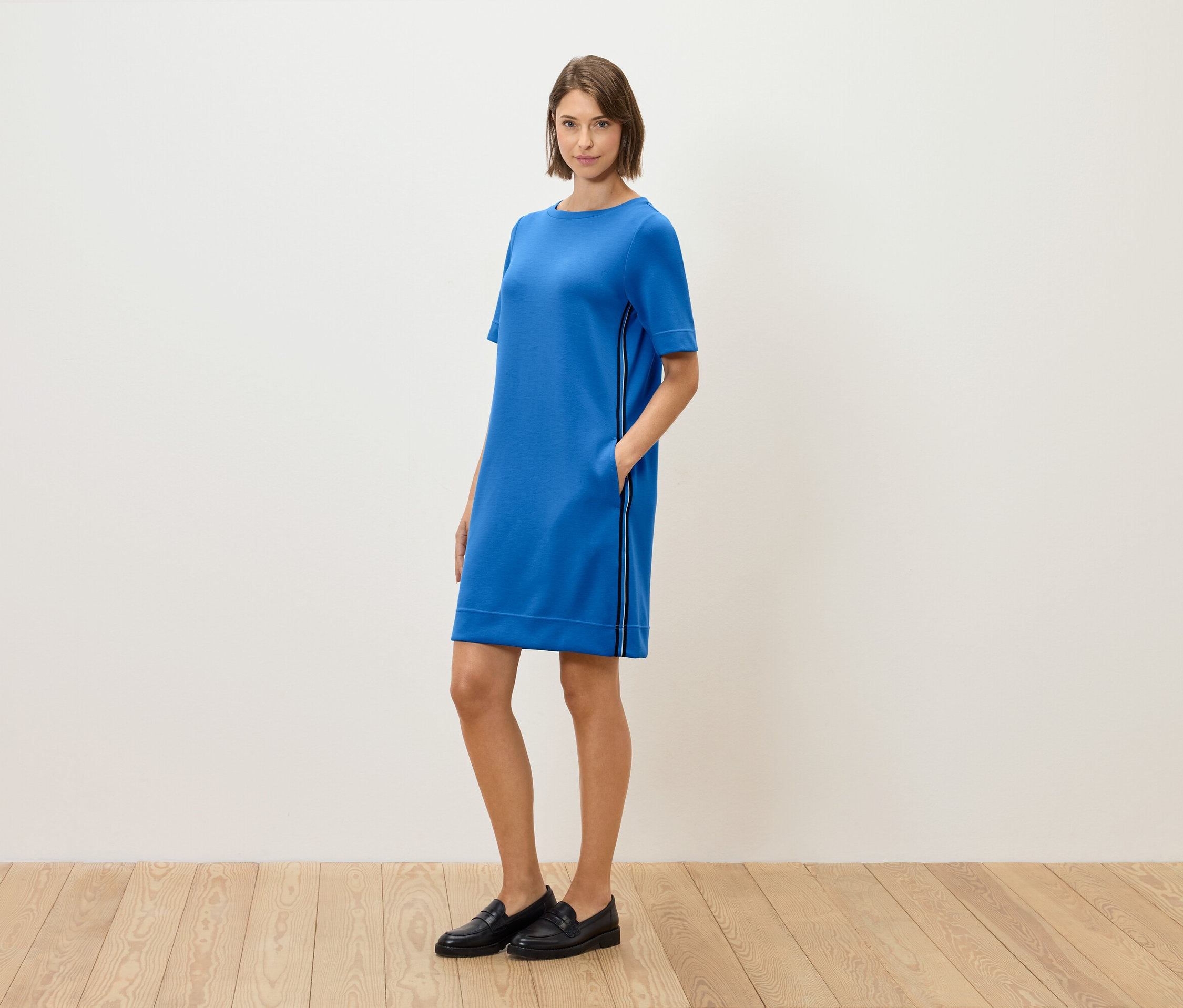 Žena stojí a pózuje v modrých šatech s krátkým rukávem a černých mokasínách.