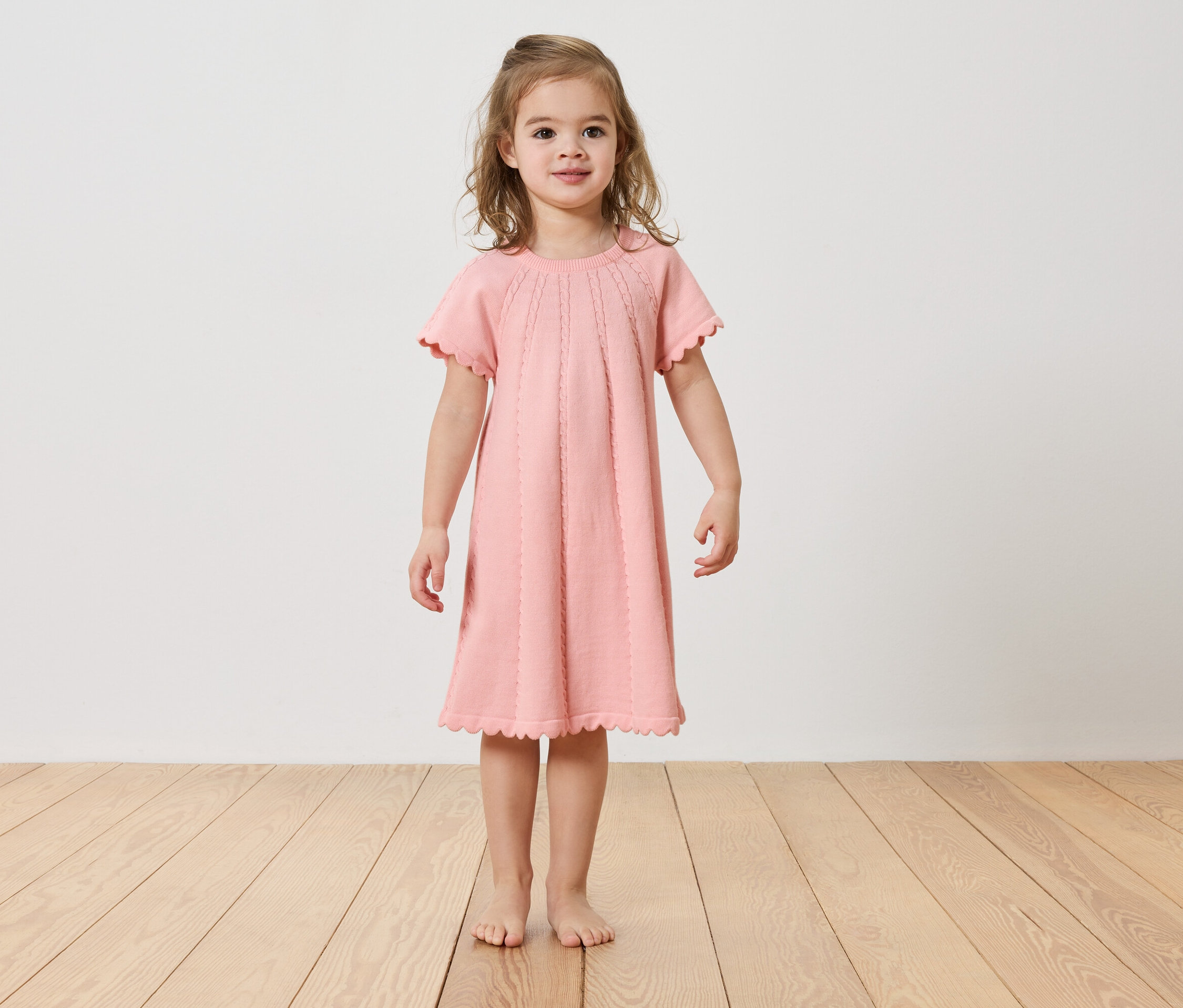 Holčička stojí bosá na dřevěné podlaze a má na sobě růžové šaty.