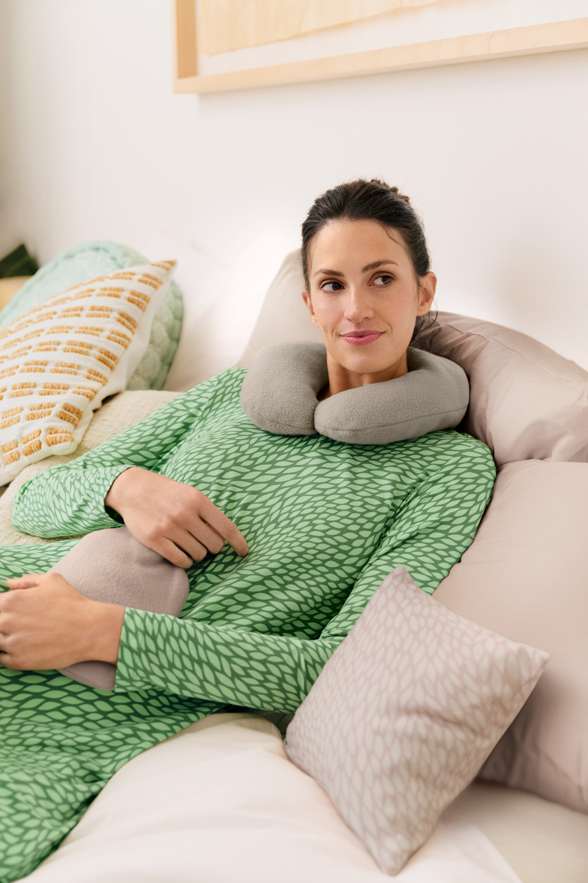 Žena leží v posteli se šedým cestovním polštářem a termoforom, oblečená v zelené noční košili.