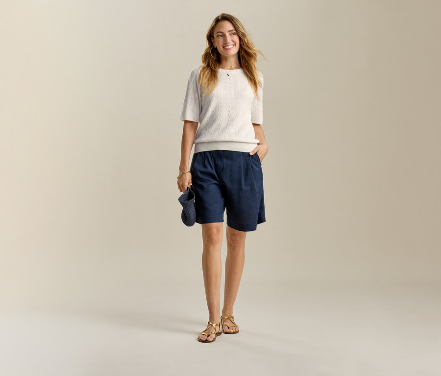 Žena ve světlém topu a tmavě modrých lněných bermudách Leinen-Bermuda-Shorts, s rukou v kapse.