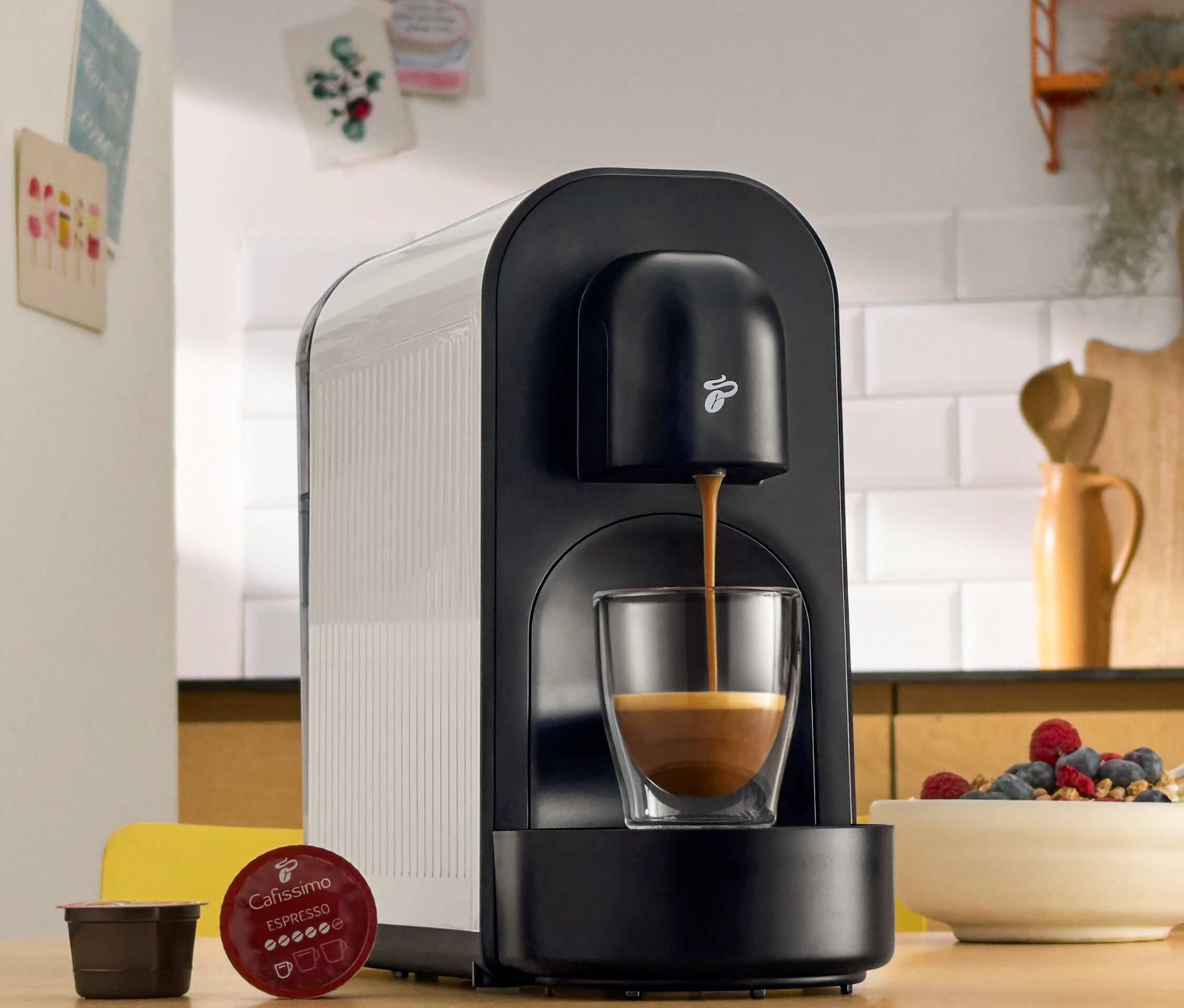 Černý kávovar nalévá kávu do sklenice. Vedle něj leží kávové kapsle a miska s bobulemi.