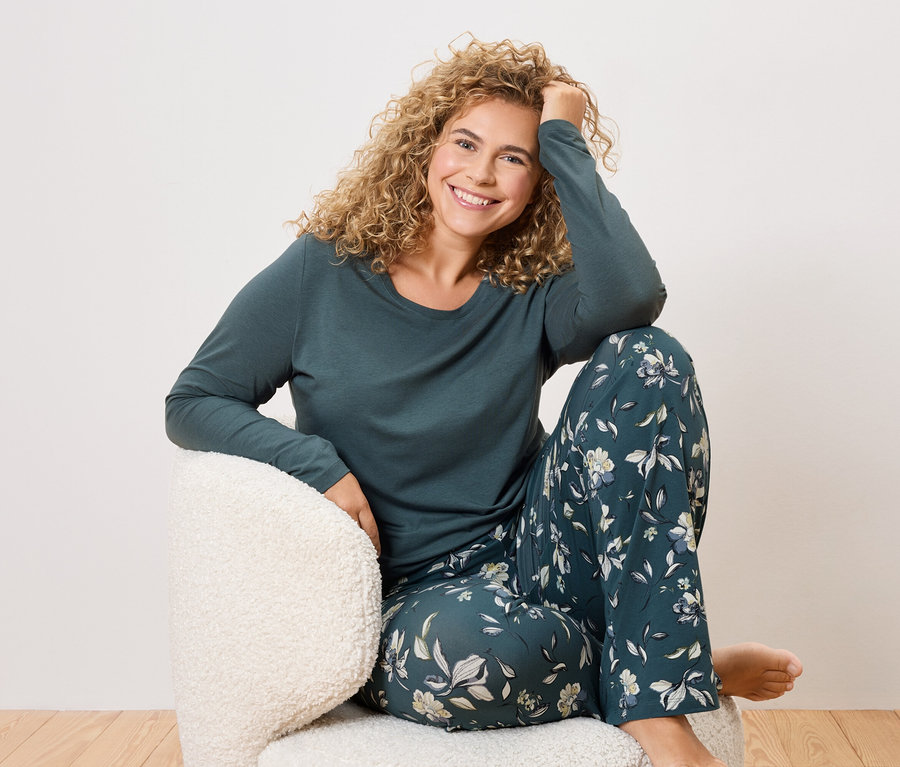 Žena s kudrnatými vlasy sedí v křesle a má na sobě tmavě zelené pyžamo s květinovým vzorem.