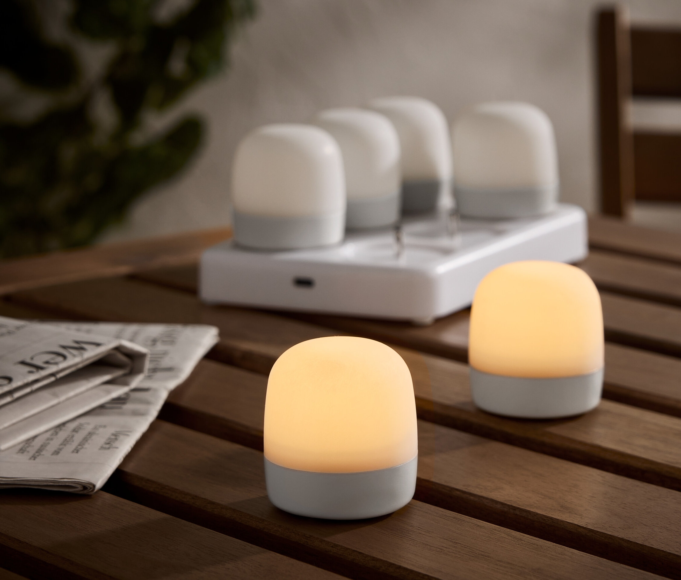 Dvě osvětlené bezdrátové stolní lampy na dřevěném stole, s nabíjecí stanicí a čtyřmi dalšími lampami v pozadí. Vedle jsou noviny.