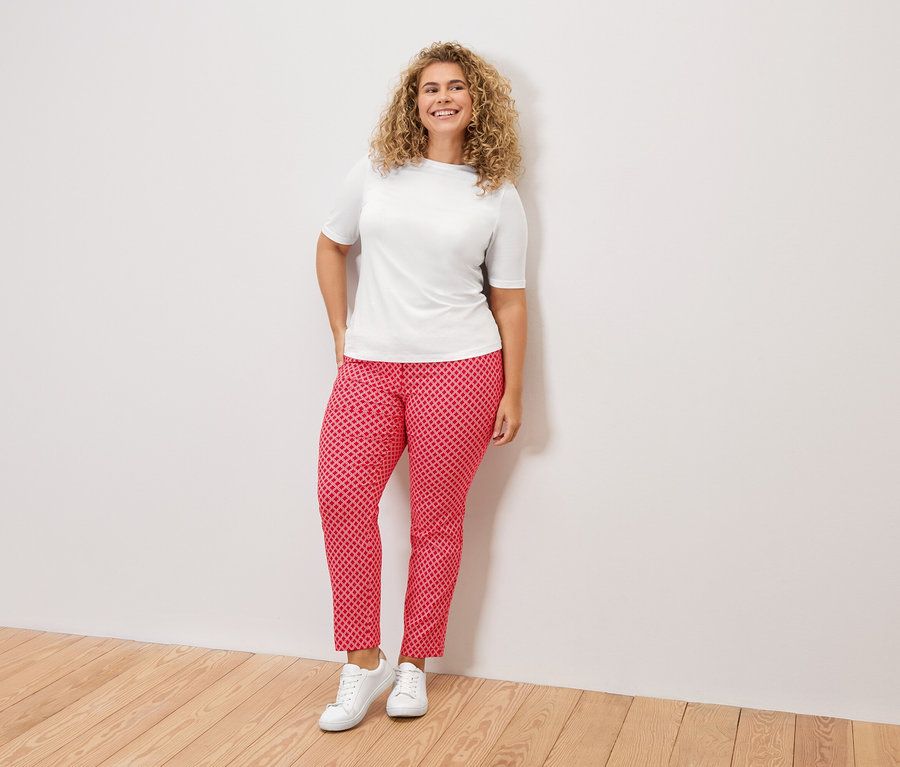 Žena s kudrnatými vlasy v bílém tričku, červených vzorovaných kalhotách a bílých teniskách stojí před bílou zdí.