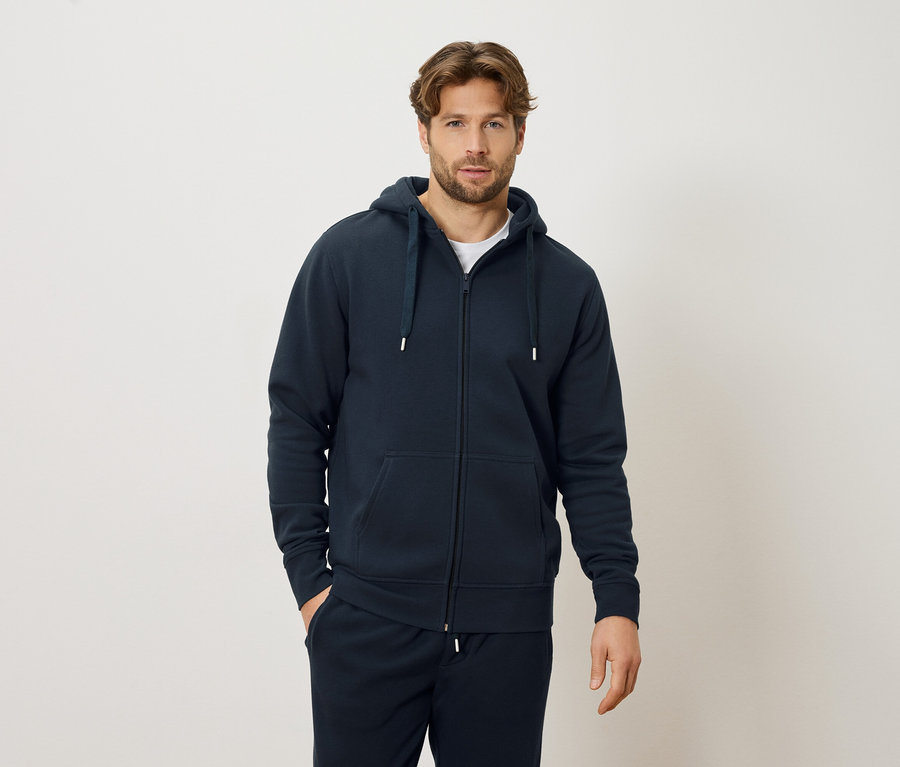 Muž v tmavě modré mikině na zip a tmavě modrých teplákách, stojící na krémovém pozadí.