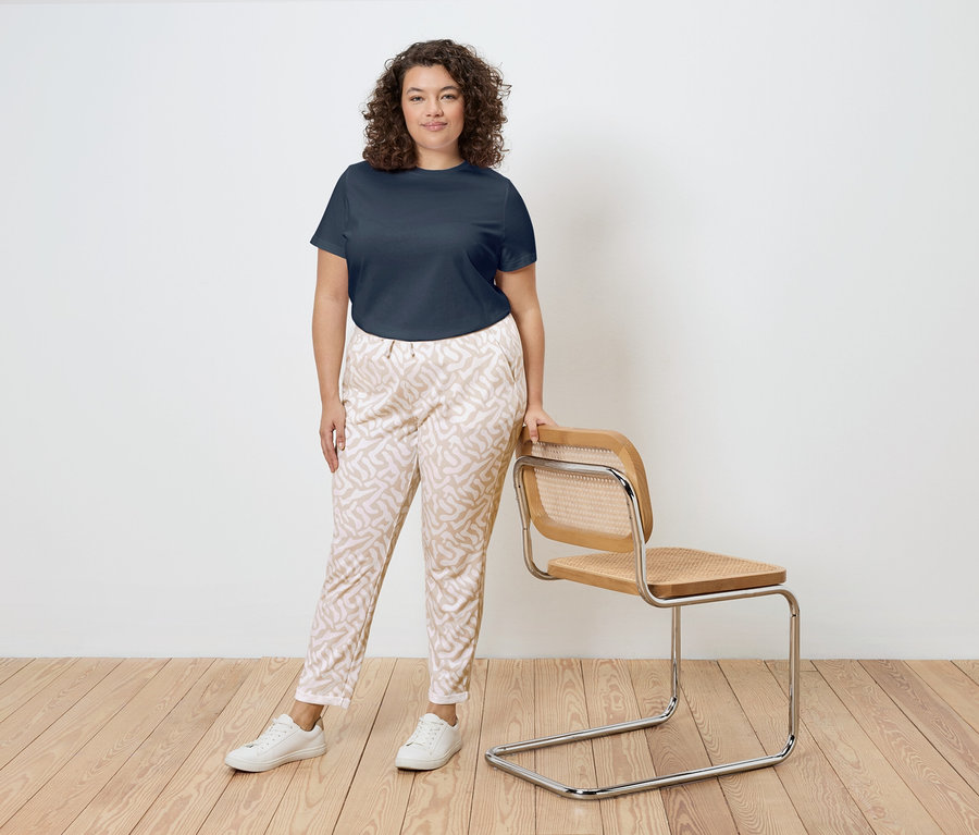 Žena s kudrnatými vlasy stojí vedle židle s dřevěným rámem a ratanovým sedákem. Má na sobě tmavě modré tričko, vzorované kalhoty a bílé tenisky.