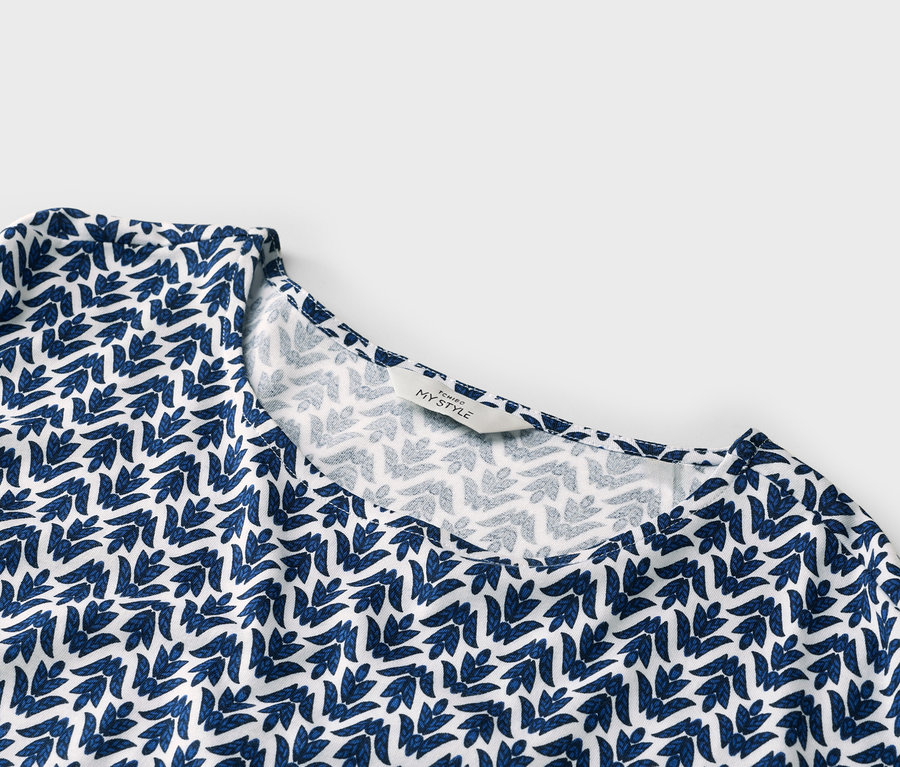Detail bílého topu s modrým listovým vzorem a štítkem s nápisem „TCHIBO MY STYLE“.