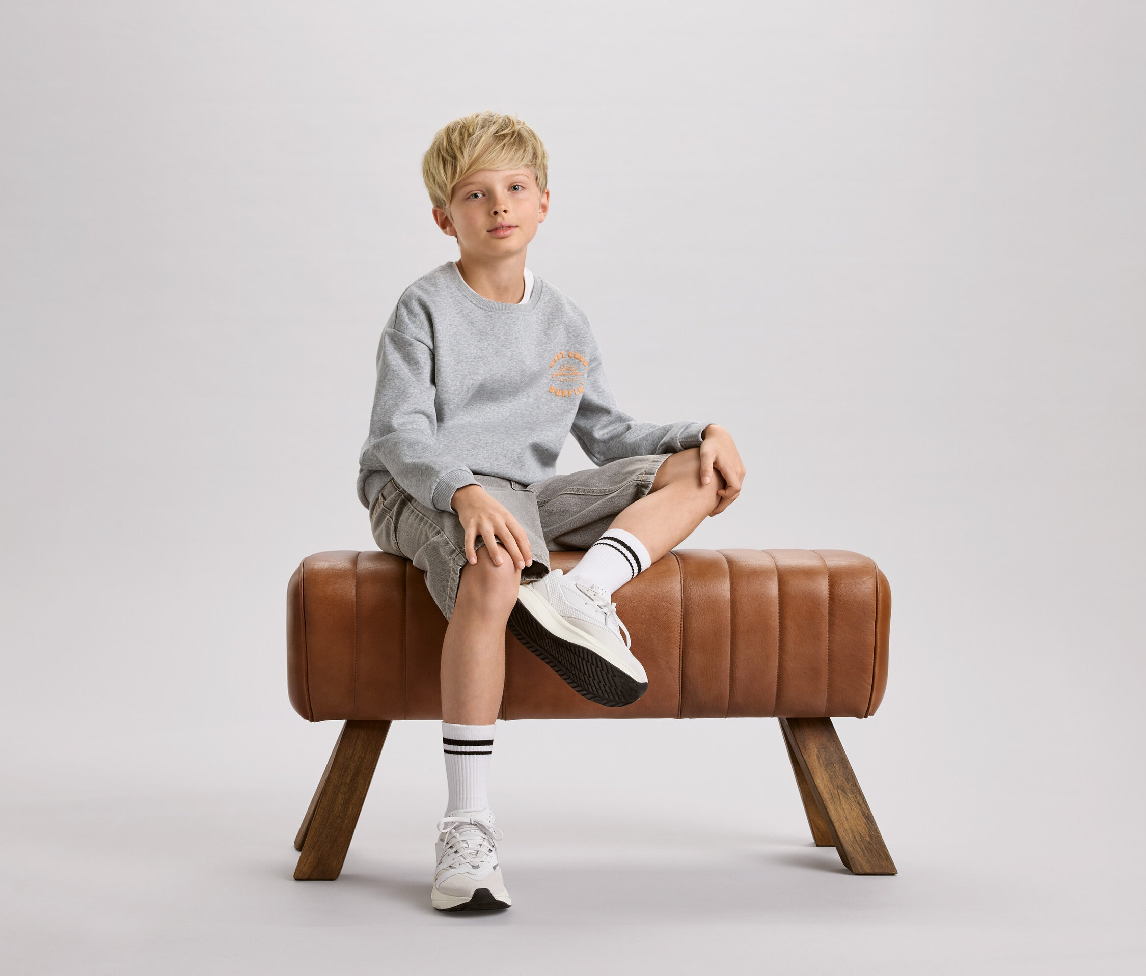 Blonďatý chlapec sedí na hnědém gymnastickém koni. Má na sobě šedou mikinu, kraťasy a bílé ponožky s černými pruhy.