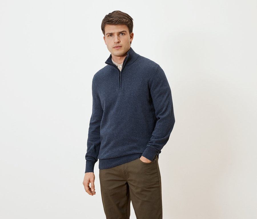 Muž v modrém svetru na zip a hnědých kalhotách s rukou v kapse na neutrálním pozadí.