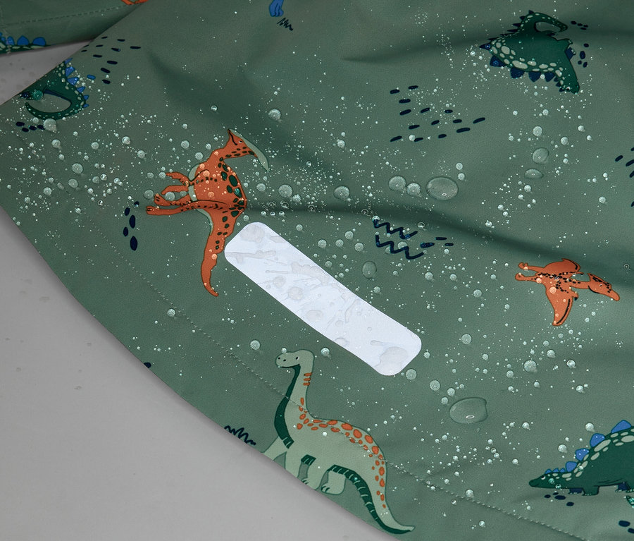 Detailní záběr na zelenou látku se vzorem dinosaurů a kapkami vody, s reflexní, obdélníkovou nášivkou.