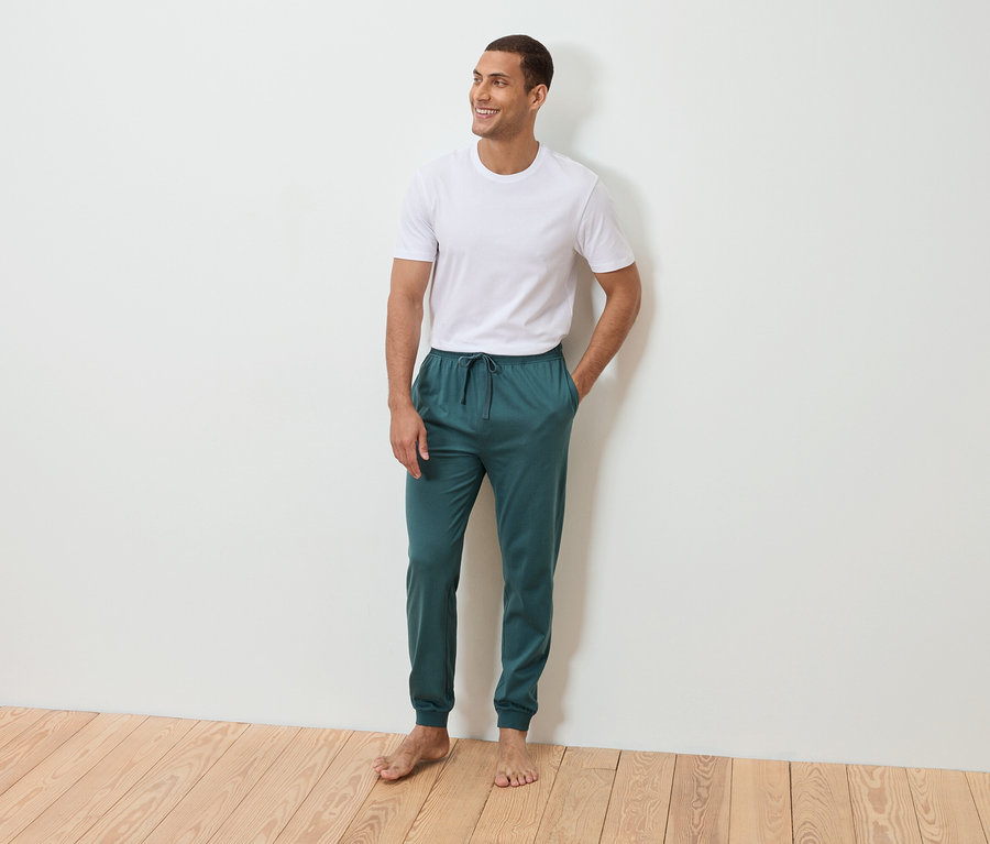 Muž stojí bosý s rukou v kapse před bílou zdí. Má na sobě bílé tričko a petrolejové tepláky.