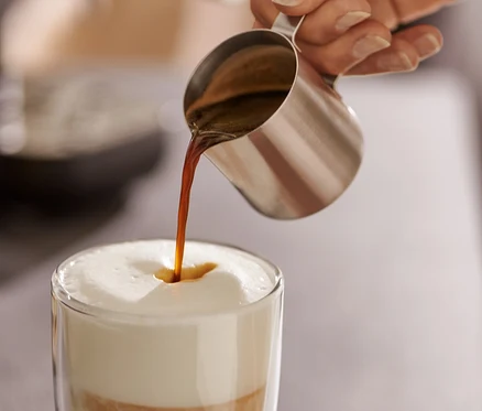 Žena nalévá espresso z konvičky z ušlechtilé oceli (80 ml) do sklenice s mlékem.