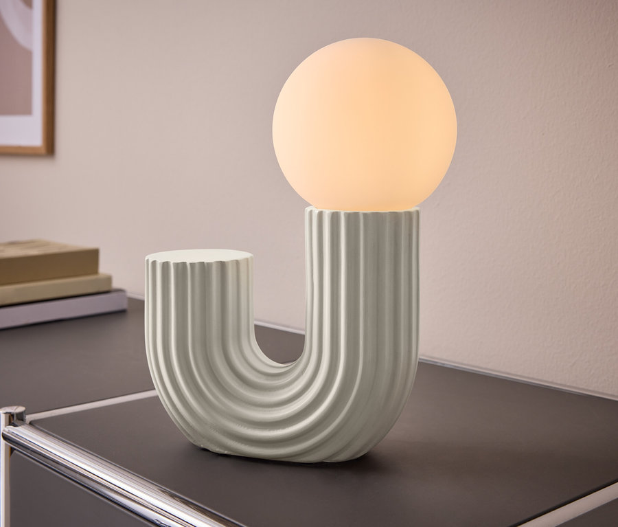 Stolní lampa ve tvaru U s žebrovanou základnou a kulovou svítící žárovkou, umístěná na tmavé skříňce. V pozadí jsou knihy a obraz na zdi.