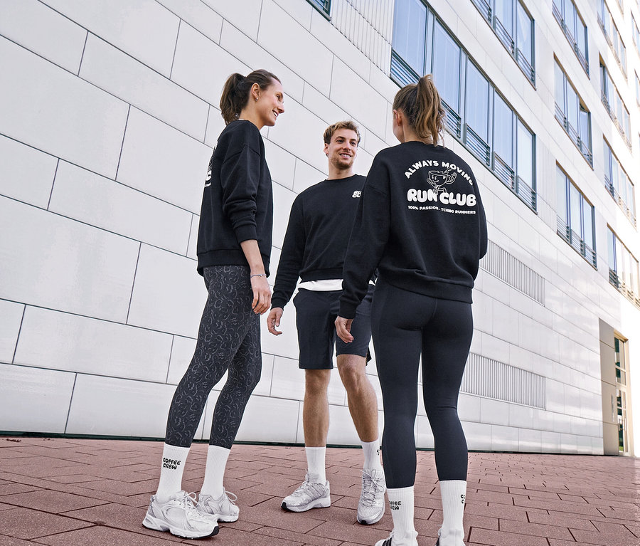 Tři lidé v černých mikinách a sportovním oblečení si povídají. Žena má na sobě šedé/černé sportovní legíny »ActivePower« a unisex tenisové ponožky Coffee Crew. Muž má na sobě černé sportovní šortky a bílé ponožky. Druhá žena má na sobě černé sportovní legíny »ActivePower« a mikinu s logem „RUN CLUB“.