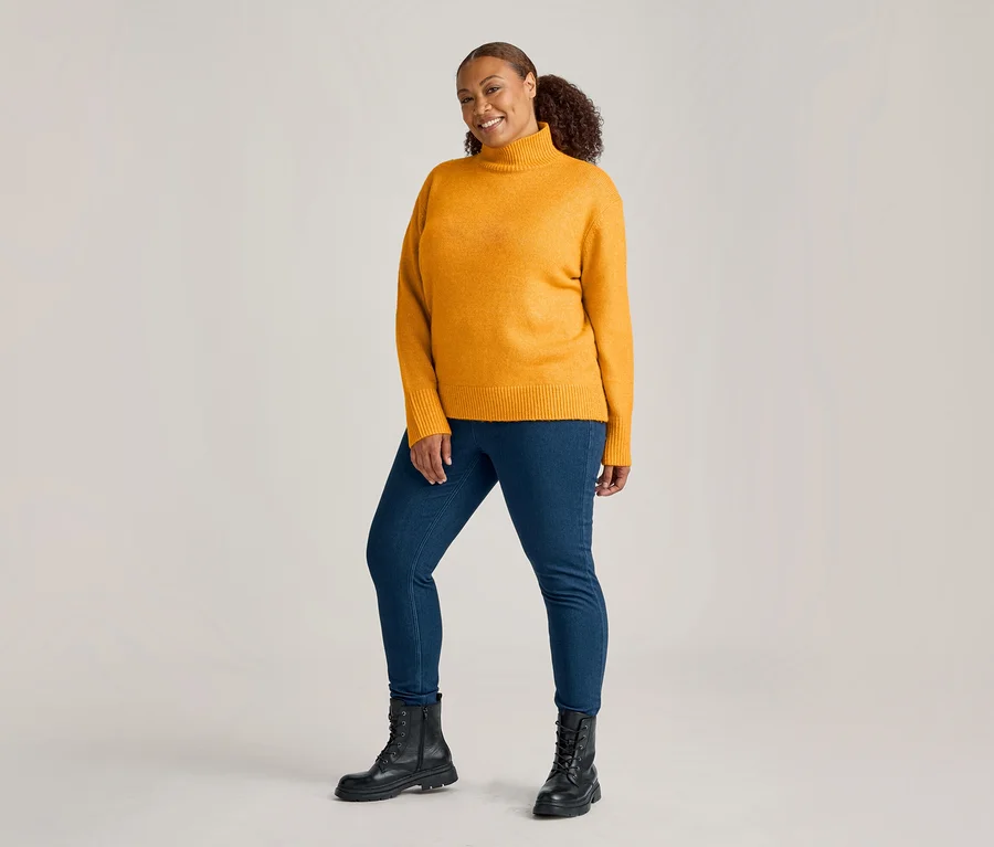 Žena v hořčicově žlutém roláku, džegínách mid blue denim a černých šněrovacích botách se usmívá do kamery.