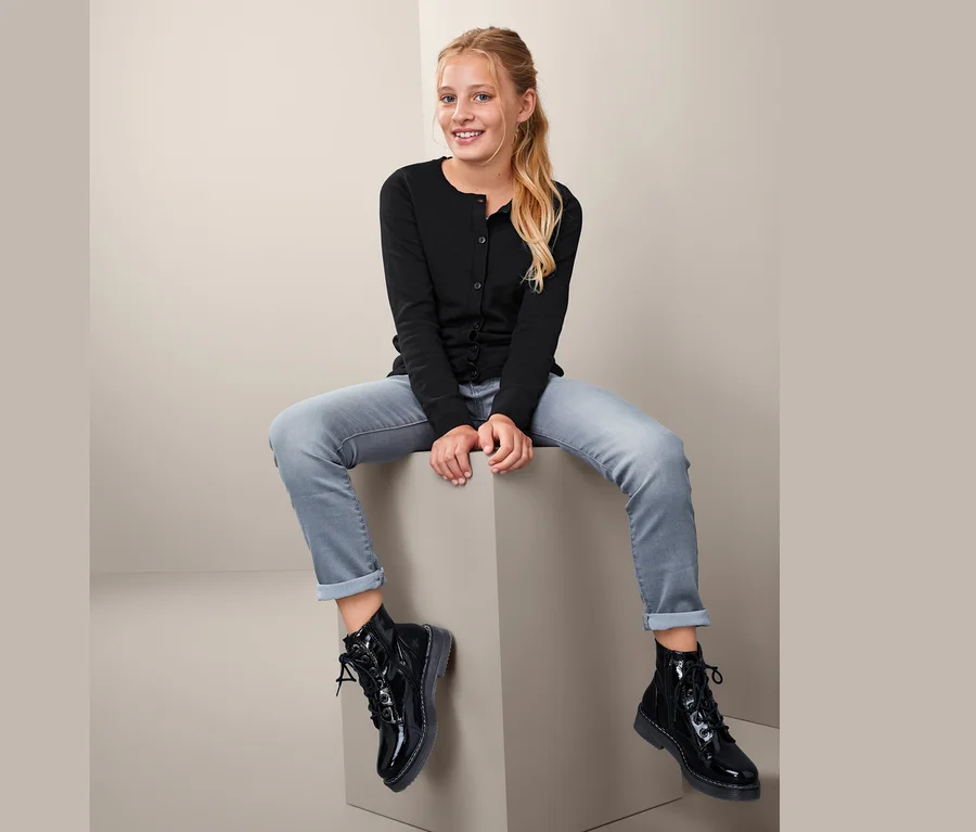 Dívka sedí na kostce a má na sobě černý kabátek z jemné pleteniny, džíny a černé boty.