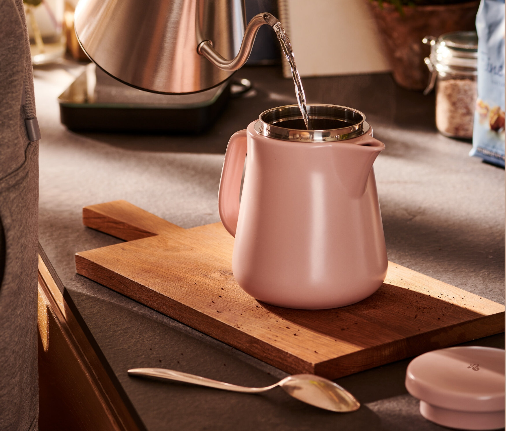 Žena nalévá vodu z nerezové konvice do keramické konvice na přípravu kávy růžové barvy.