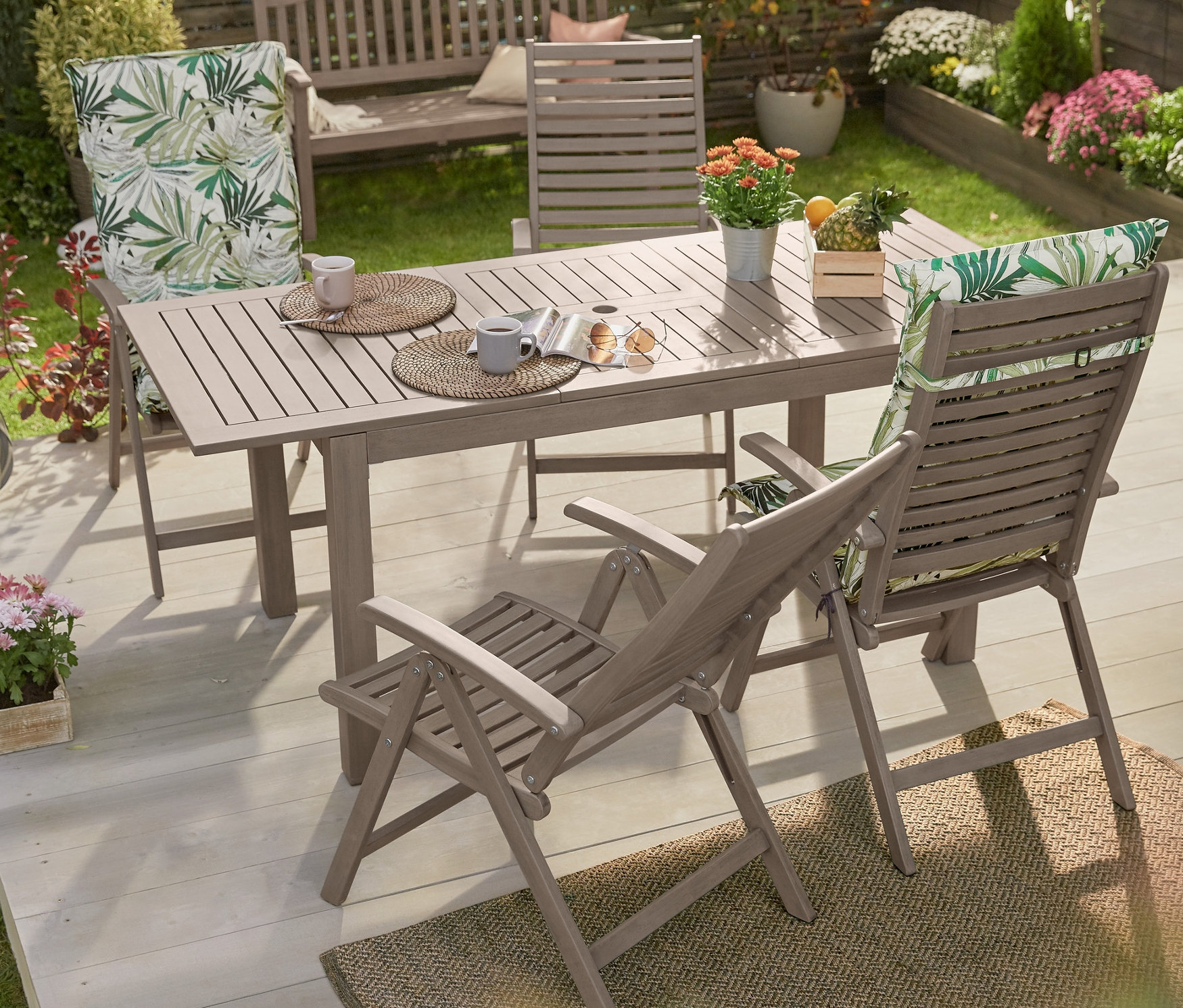 Rozkládací zahradní stůl, vintage grey, obklopený židlemi s vysokým opěradlem s potiskem a zahradní lavičkou, vintage grey, na dřevěné terase.