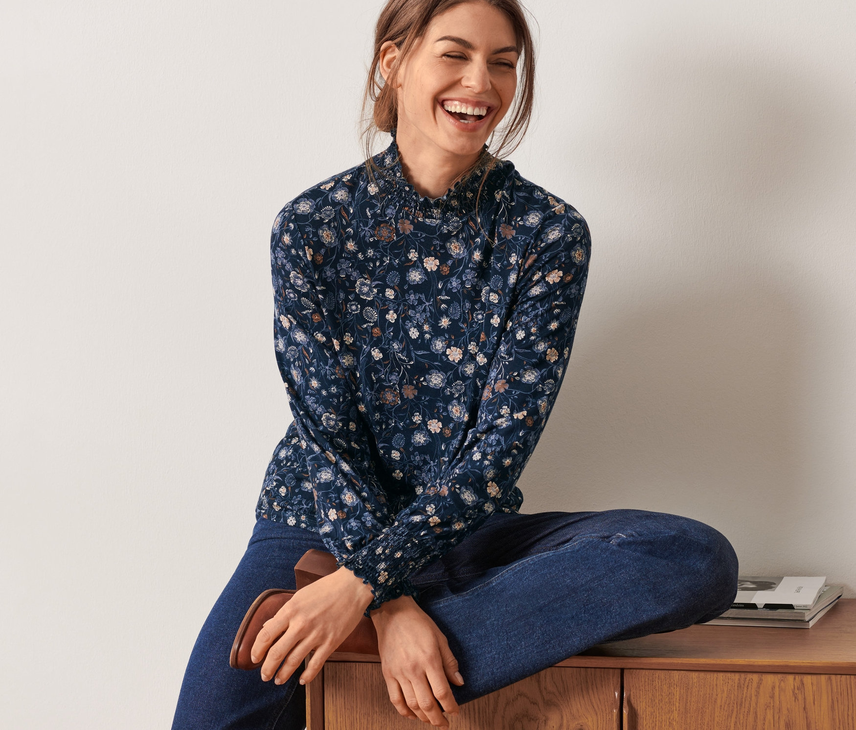 Žena sedí na dřevěné komodě a má na sobě květované halenkové triko se stojáčkem.