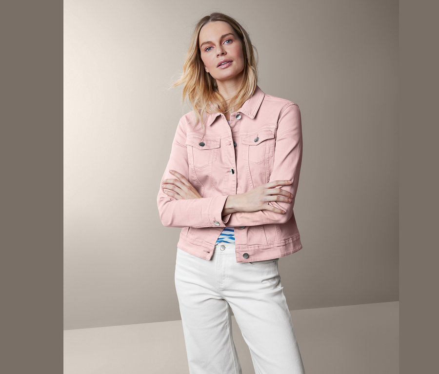 Žena pózuje v růžové džínové bundě a bílých kalhotách.