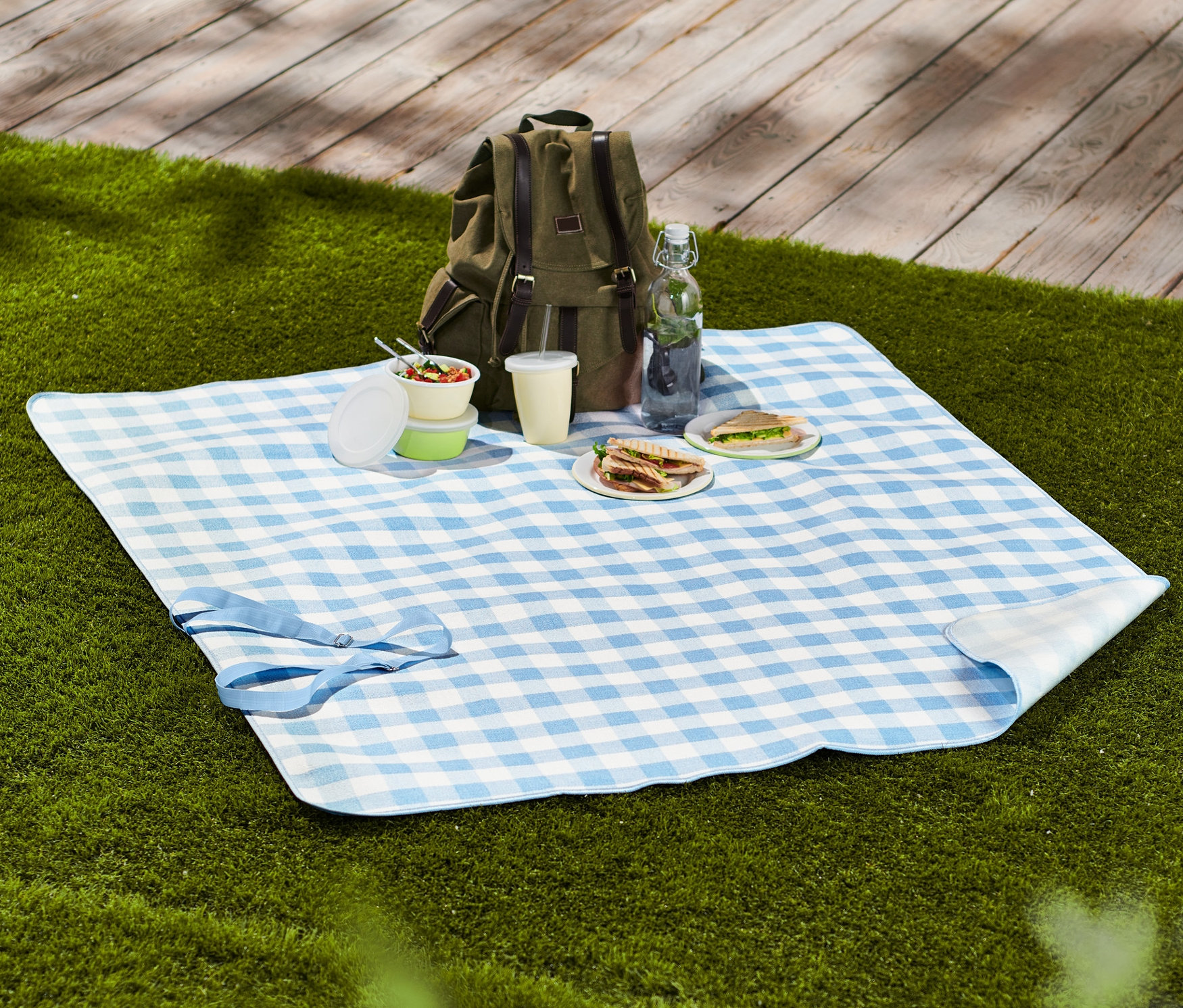 Na trávě leží malá kostkovaná pikniková deka s jídlem, pitím a batohem.