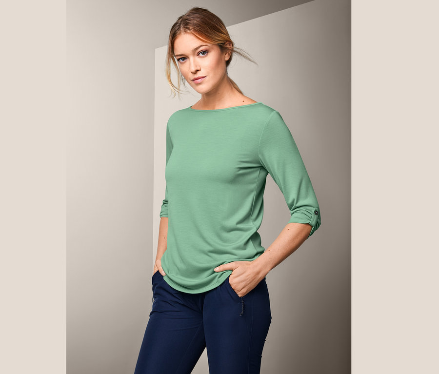 Žena pózuje v mátově zeleném funkčním tričku s 3/4 rukávy.