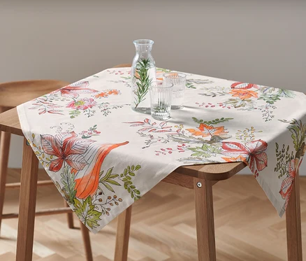Na dřevěném stole leží ubrus o rozměrech cca 90 x 90 cm s květinovým vzorem. Na ubrusu stojí karafa s vodou a dvě sklenice.
