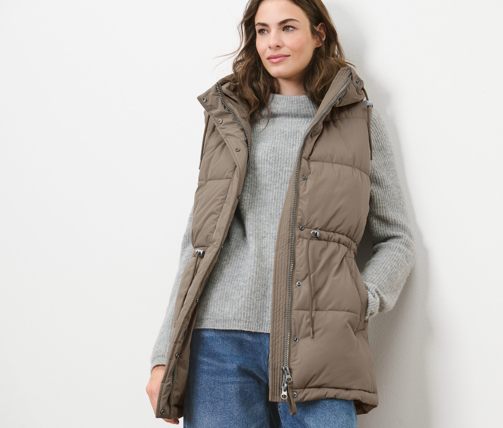 Žena má na sobě prošívanou vestu s kapucí, svetr a džíny.