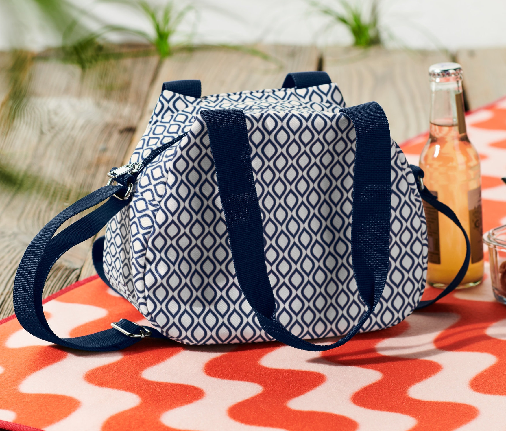 Modrobílá minichladicí taška leží vedle nápoje a misky s těstovinami na oranžovo-bílém vlnitém plážovém ručníku.