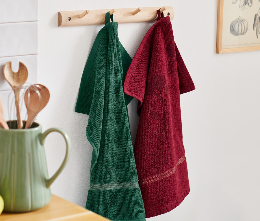 Zelený a červený froté kuchyňský ručník visí na dřevěném věšáku vedle zeleného džbánu s kuchyňským náčiním.
