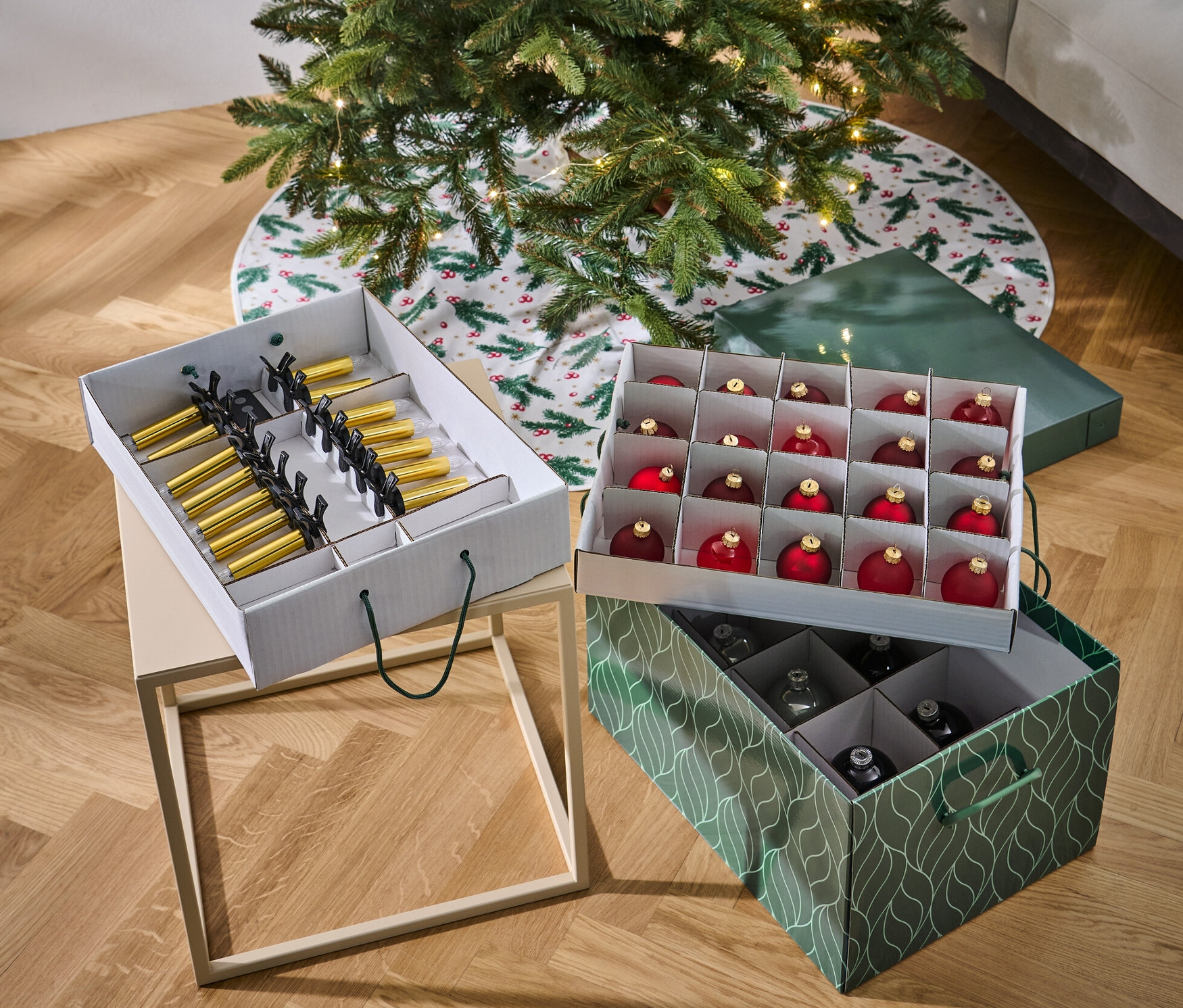 Vánoční stromek stojí vedle dvou boxů na vánoční ozdoby. Jeden box je bílý a obsahuje zlaté a černé ozdoby, druhý je zelený a obsahuje červené a stříbrné ozdoby.