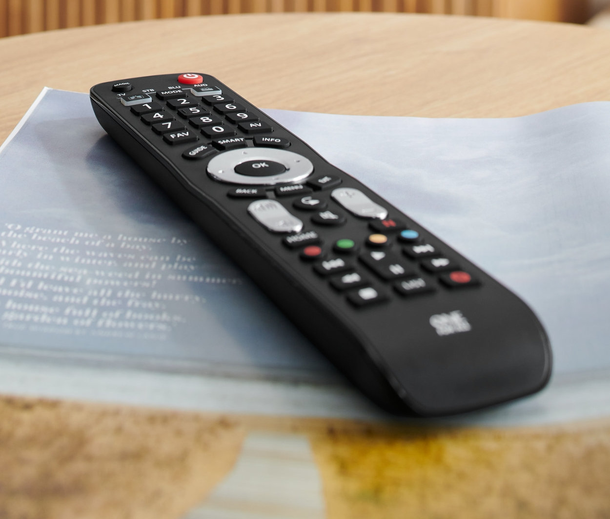 Černý univerzální dálkový ovladač 4 v 1 leží na stole vedle sklenice s vodou a časopisu.