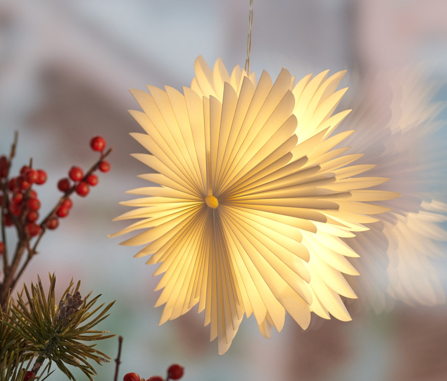 Zavěšená malá papírová hvězda s LED diodami u okna, vedle větví s červenými bobulemi.