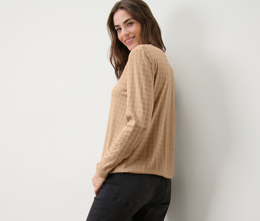 Usmívající se žena se dívá přes rameno a pózuje v pískovém strukturovaném triku s dlouhým rukávem a černých džínách.