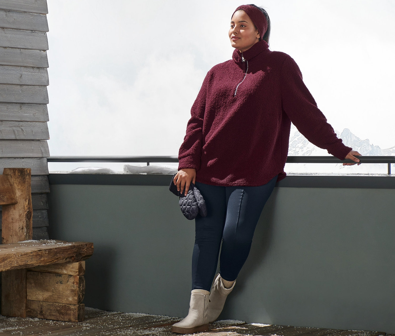 Žena opřená o zábradlí má na sobě plyšovou mikinu oversized, pletenou čelenku, modré termolegíny a kotníkové boty z velurové kůže. V ruce drží prošívané palčáky.