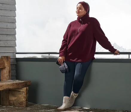 Žena opřená o zábradlí má na sobě plyšovou mikinu oversized, pletenou čelenku, modré termolegíny a kotníkové boty z velurové kůže. V ruce drží prošívané palčáky.