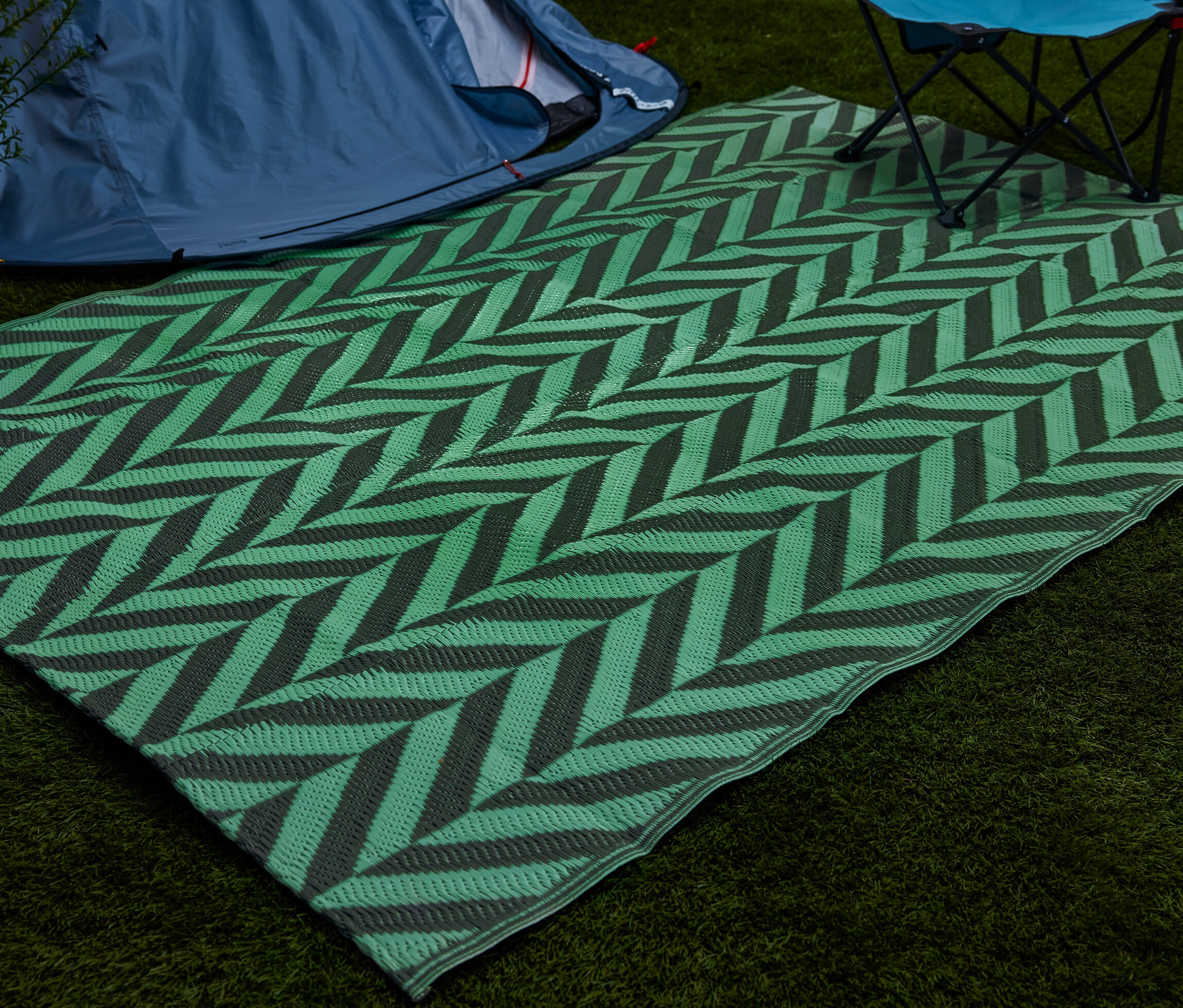 Zelený fluorescenční stanový koberec s šedým vzorem chevron leží na trávě vedle stanu a skládací židle.