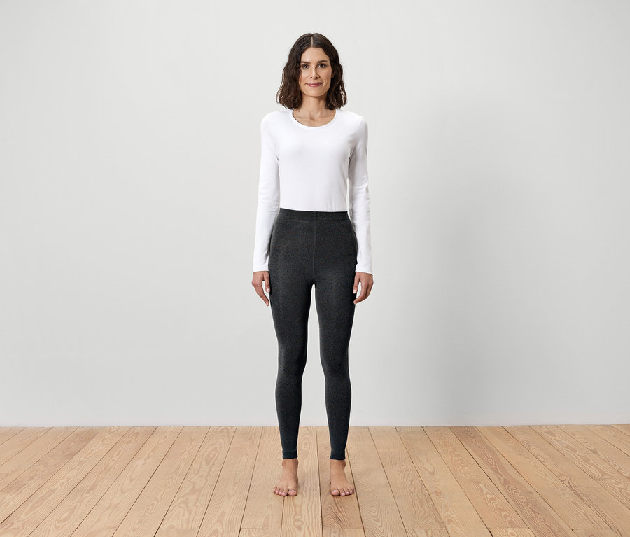 Žena stojí naboso na dřevěné podlaze a má na sobě bílé tričko s dlouhým rukávem a šedé legíny.