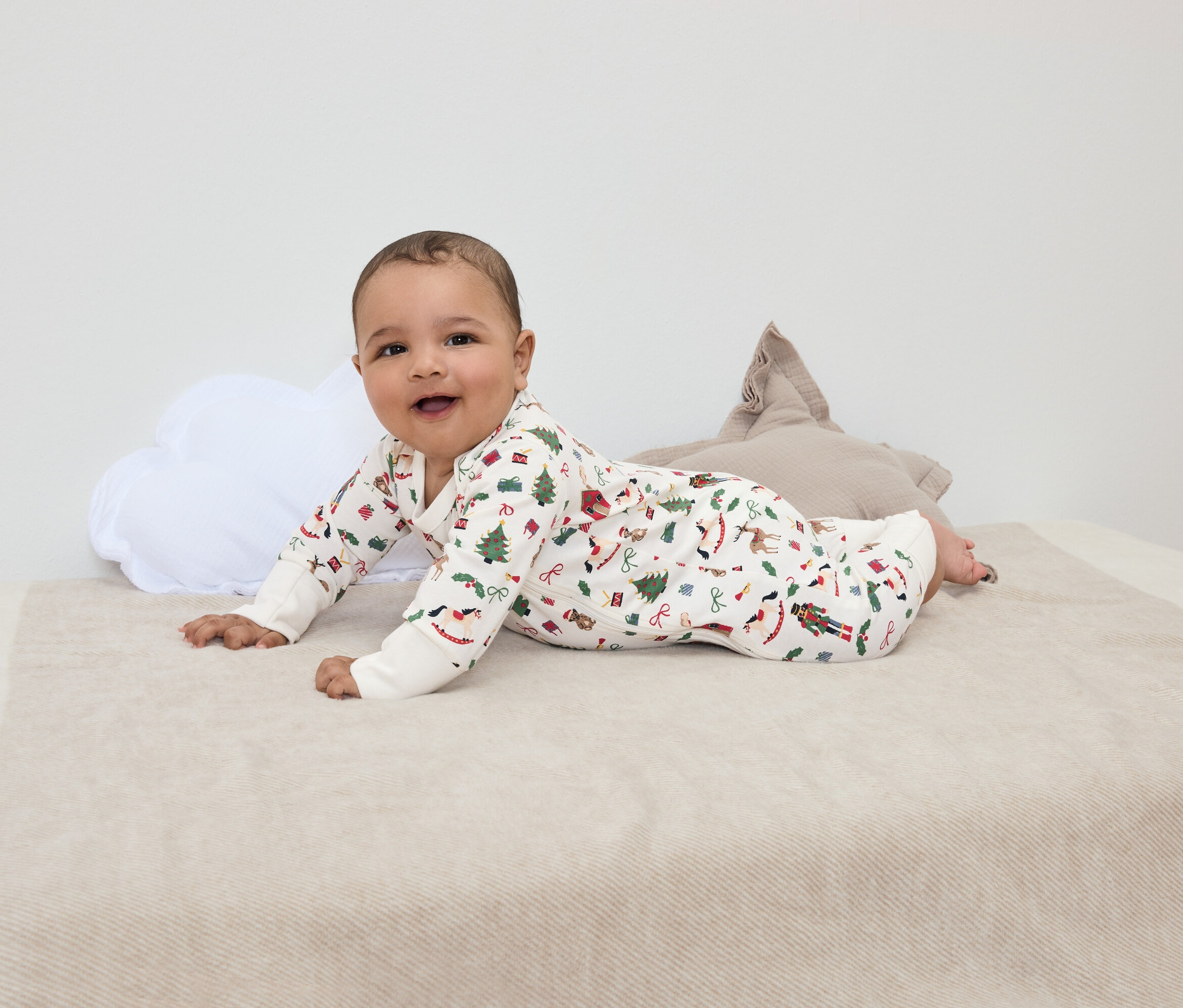 Usmívající se kojenec leží na břiše na dece s vánočním potiskem, vedle bílého polštáře ve tvaru mraku a hnědého hvězdného polštáře.