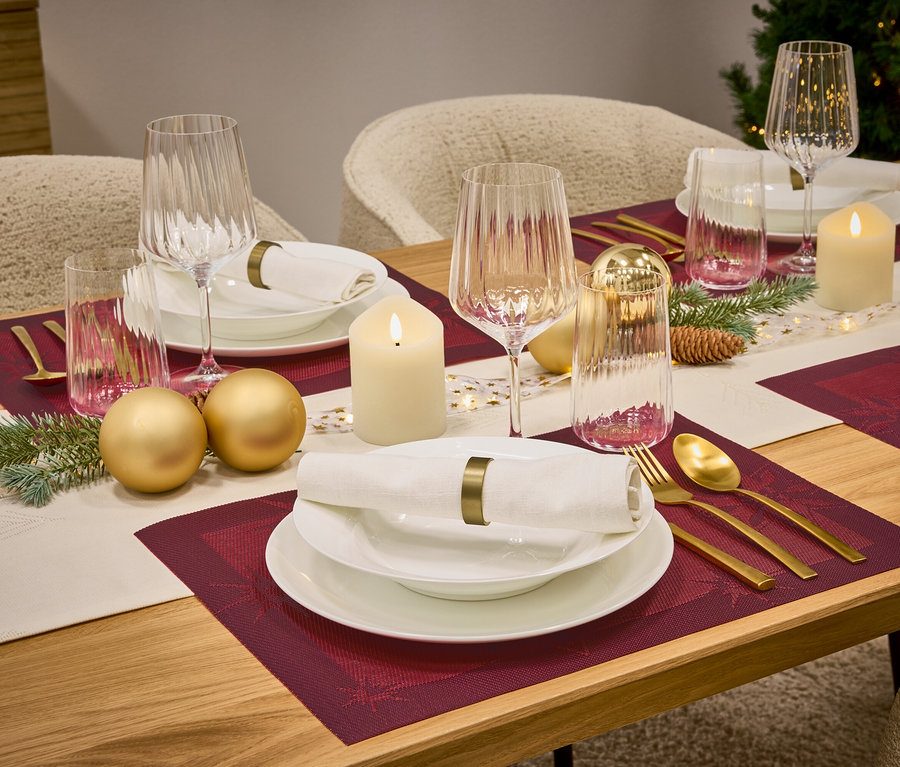 Prostřený stůl s vínovými prostíránímí, bílým nádobím, sklenicemi, zlatými příbory, svíčkami a ozdobami.