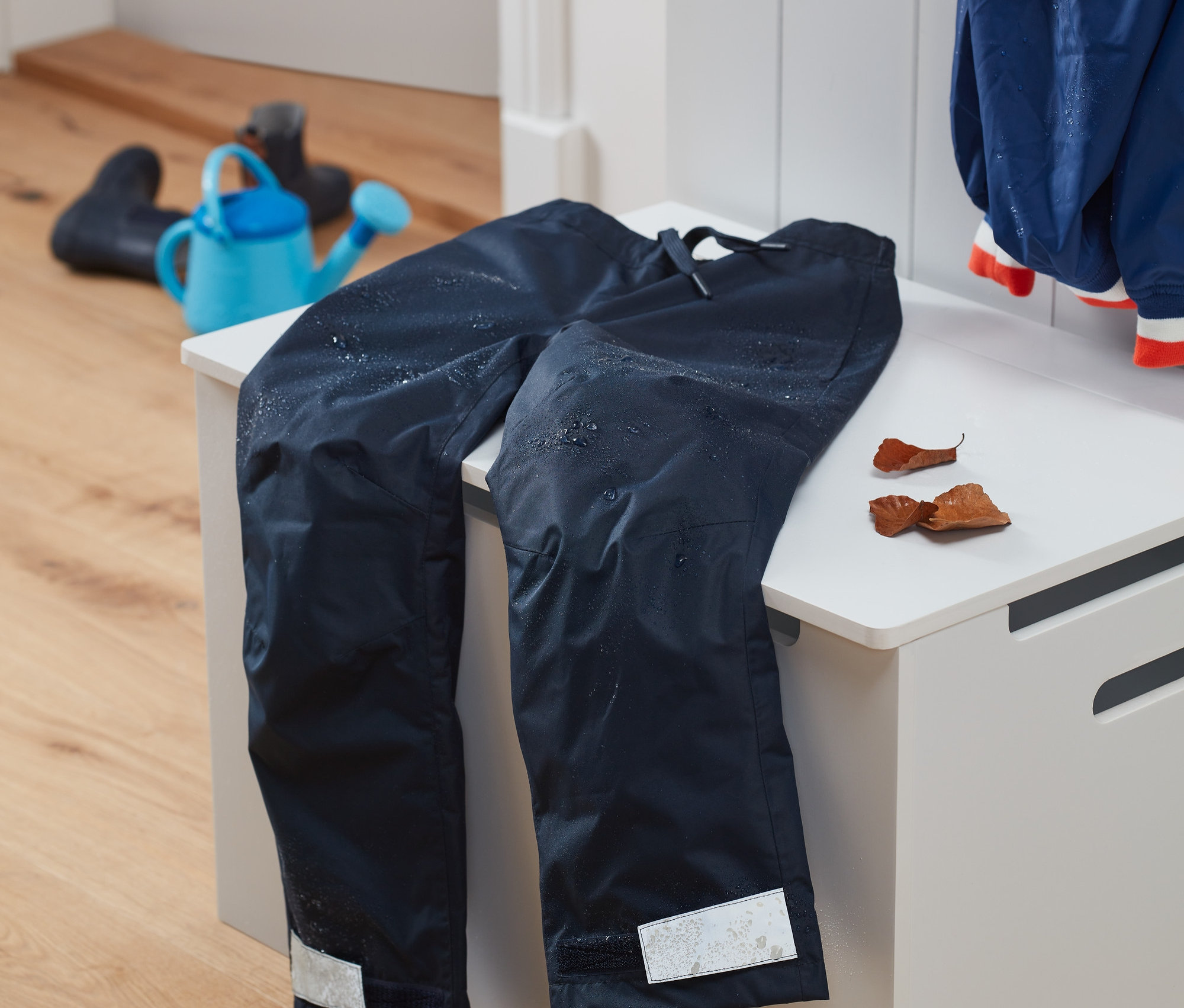 Tmavě modré kalhoty do deště z recyklovaného materiálu leží na bílé lavici. V pozadí je modrá konev a holínky.