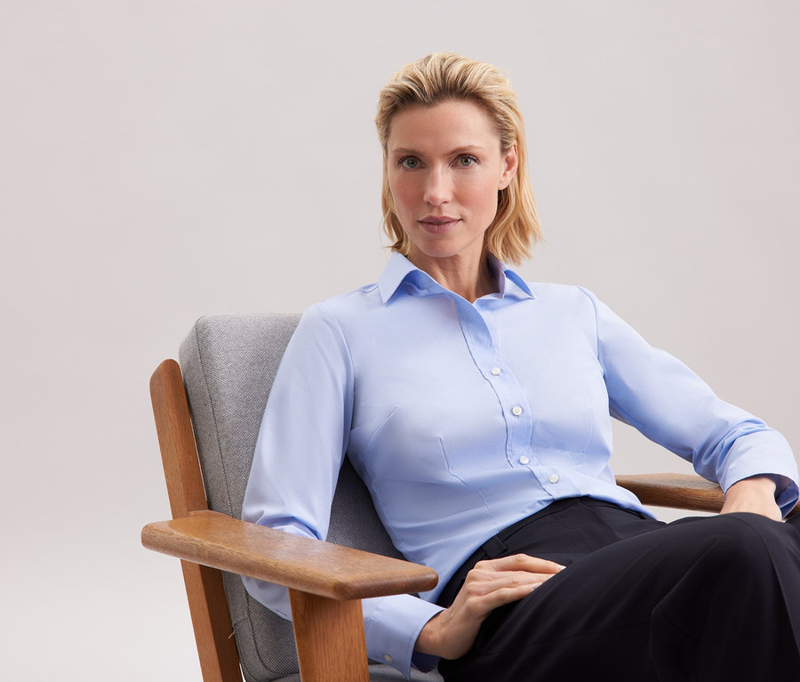 Blondýna sedí v šedém křesle a má na sobě modrou košili a černé kalhoty.