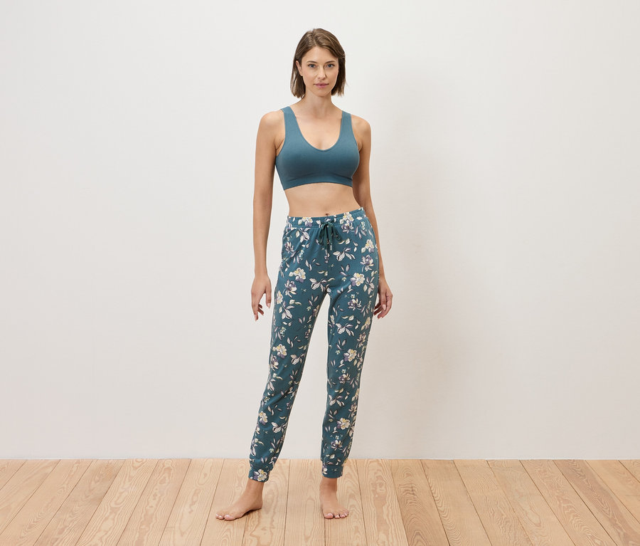 Žena stojí bosá před zdí a má na sobě modrou sportovní podprsenku a modré tepláky s květinovým vzorem.