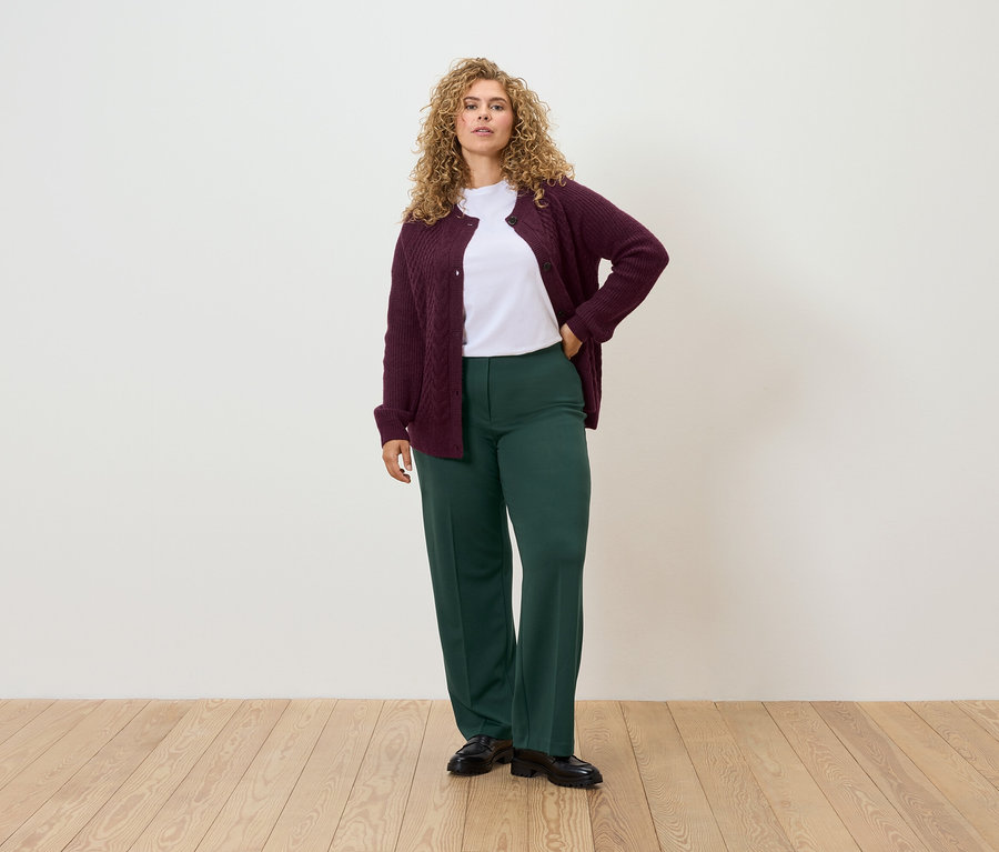 Žena stojí a má ruku na boku, oblečená v bordó kardiganu, bílém tričku a zelených kalhotách.