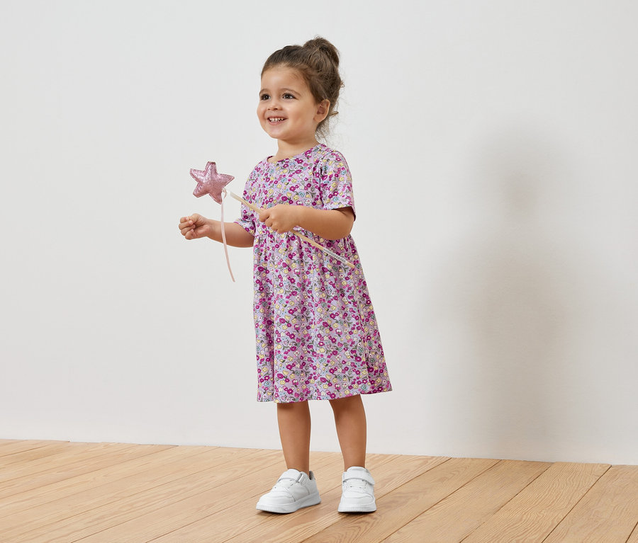 Usmívající se holčička drží hvězdnou hůlku a má na sobě květinové šaty a tenisky.